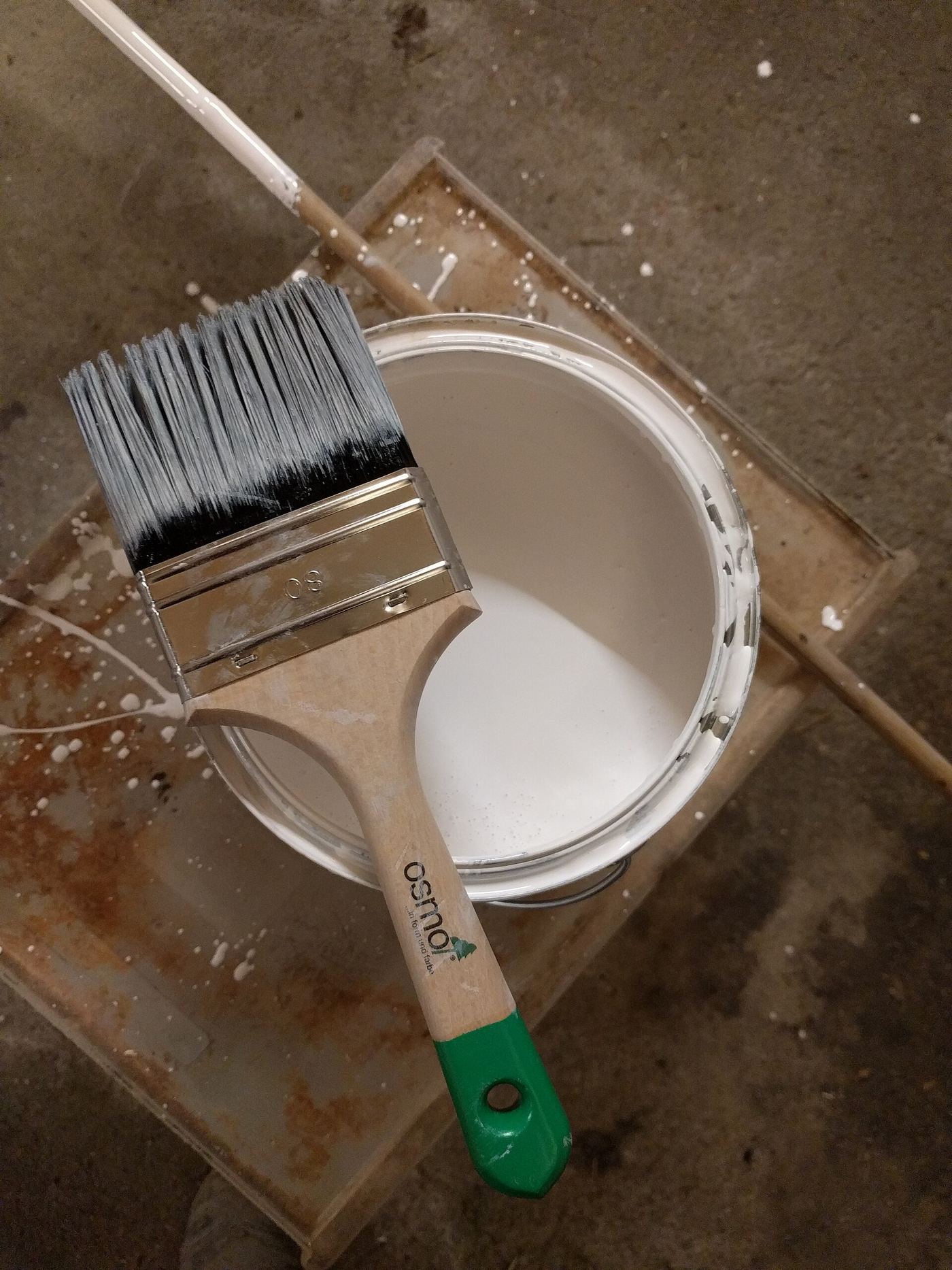 Open pot witte verf met een brede kwast erop, op een bevuilde werkplank