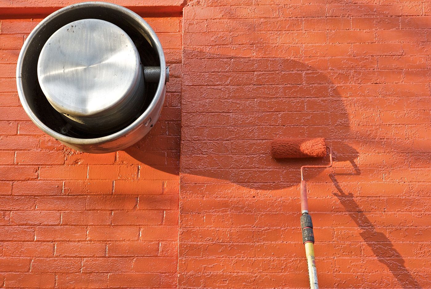 Verfrol met een lange steel schildert een bakstenen muur oranje, naast een metalen afvoer