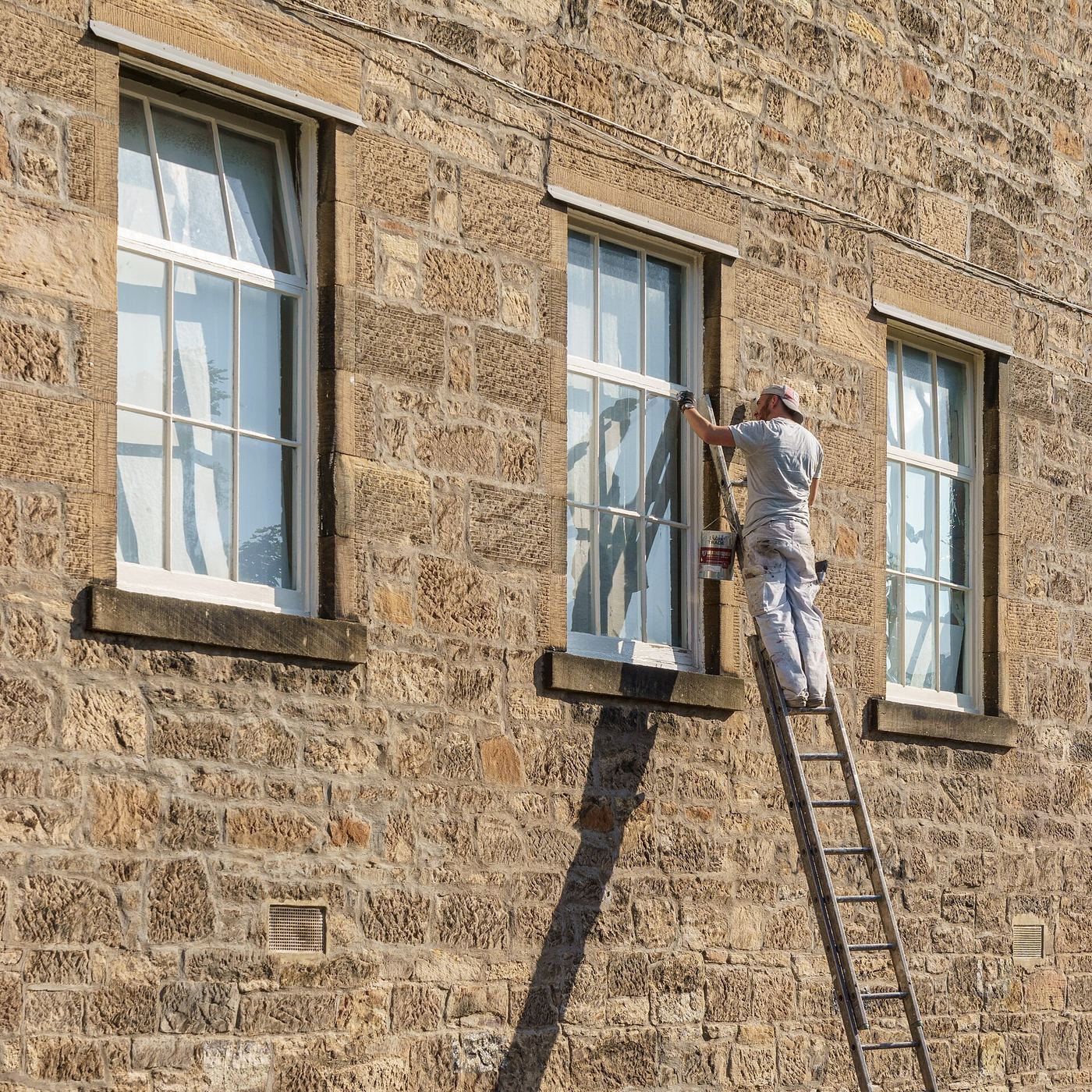 Schilder op een ladder schildert de kozijnen van een raam in een gevel van natuursteen
